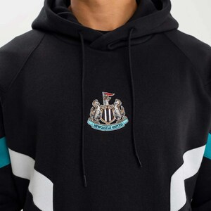 May include: Black hoodie with a hood and drawstring. The front features a Newcastle United crest with a red flag. Teal and white accents are on the sleeves. The hoodie is made of a soft, comfortable material.