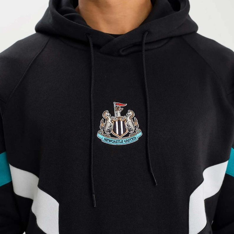 May include: Black hoodie with a hood and drawstring. The front features a Newcastle United crest with a red flag. Teal and white accents are on the sleeves. The hoodie is made of a soft, comfortable material.
