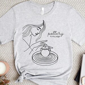 May include: A light gray t-shirt featuring a line drawing of a person working on a pottery wheel. The text "pottery is my yoga" is printed in a script font. The shirt is styled with a knot at the waist.