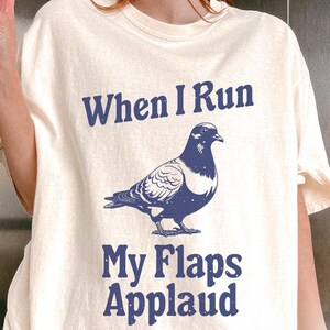 May include: A white t-shirt with a blue graphic of a pigeon and the text "When I Run My Flaps Applaud". The shirt is labeled "Ivory".