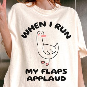 May include: A white t-shirt with the text "When I run my flaps applaud" and a cartoon illustration of a goose. The shirt is available in the color ivory.
