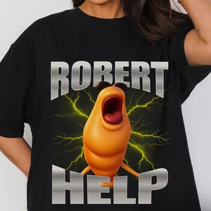 May include: Black t-shirt with a cartoon character design. The design includes an orange character with an open mouth, yellow lightning bolts, and the words "ROBERT" and "HELP" in a metallic font. The word "Black" is printed at the bottom.