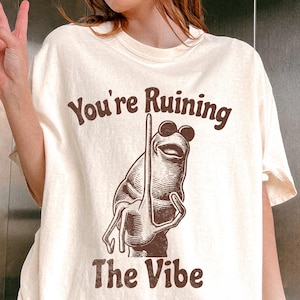 May include: Ivory t-shirt featuring the text "You're Ruining The Vibe" in brown. The design shows a cartoon frog wearing sunglasses. The shirt has a relaxed fit.