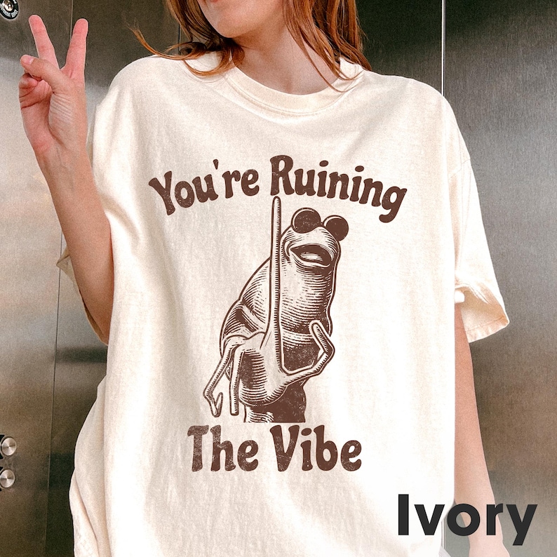 May include: Ivory-colored t-shirt with the text "You're Ruining The Vibe" in brown. The graphic features a cartoon frog wearing sunglasses. The shirt is a relaxed fit.