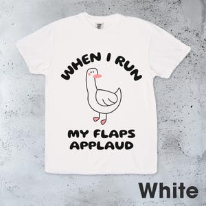 以下が含まれることがあります： 「When I run my flaps applaud」というテキストと、漫画のガチョウが描かれた白いTシャツです。