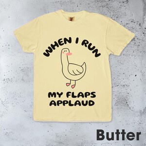 以下が含まれることがあります： 黄色いTシャツに、漫画の鴨と「When I run my flaps applaud」という文字が書かれています。文字は黒で、鴨は黄色で、足はピンク、くちばしはピンクです。
