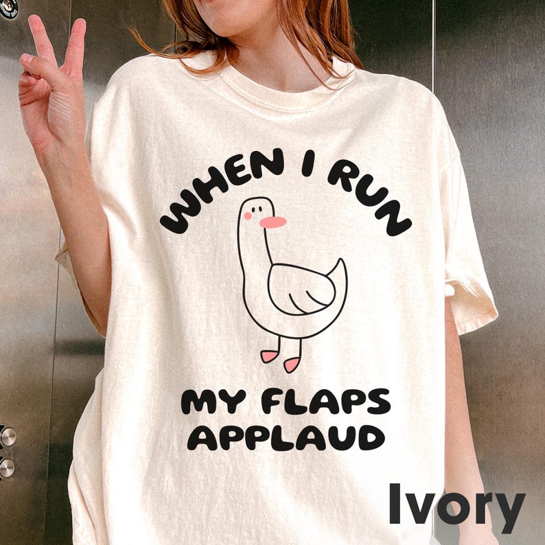 以下が含まれることがあります： 「When I run my flaps applaud」というテキストと、ガチョウの漫画イラストが描かれた白いTシャツ。アイボリーカラーで販売されています。