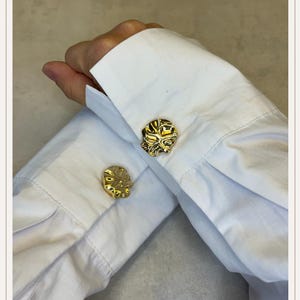 Könnte beinhalten: Nahaufnahme von goldfarbenen Manschettenknöpfen mit einem strukturierten, organischen Design. Die Manschettenknöpfe sind an den Manschetten eines weißen Hemdes befestigt. Das Bild zeigt die komplizierten Details und die glänzende Oberfläche der Manschettenknöpfe.