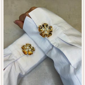 May include: Gold-tone flower-shaped cufflinks with pearl accents. The cufflinks are attached to the cuffs of a white dress shirt. The flower design has detailed petals and a central cluster of small pearls.