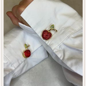 May include: Gold-coloured cufflinks shaped like a red apple, adorned with sparkling crystals. The apple features a green crystal leaf and stem. The cufflinks are shown on a white dress shirt.