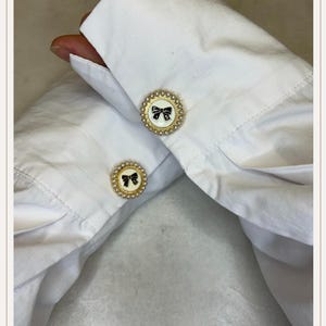 Op de afbeelding: Close-up van een witte manchet met twee decoratieve manchetknopen. Elke manchetknop heeft een goudkleurige rand met een parelachtige rand, rond een wit midden met een zwart strikdesign. Het overhemd is gemaakt van een knisperende, witte stof.