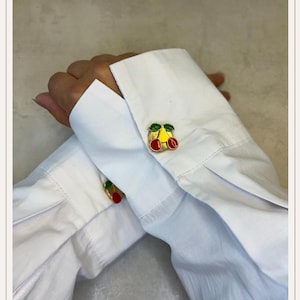 May include: Gold cufflinks featuring a cherry design. The cherries are red with green leaves and yellow detailing. The cufflinks are on a white shirt.