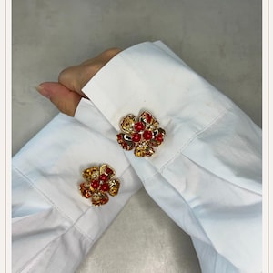 May include: Gold-toned flower-shaped cufflinks with red and amber-colored gemstones. The cufflinks are on the white cuff of a long-sleeved shirt. The flower design features a central cluster of red beads and amber-colored petals.