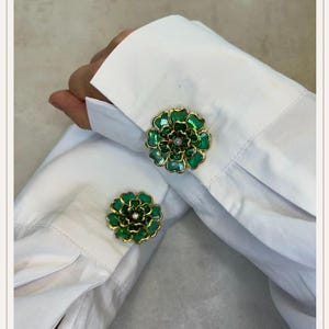 May include: Green and gold flower-shaped cufflinks with a small diamond center. The cufflinks are set against a white shirt cuff. The flower design has a detailed, layered appearance.