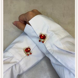 Peut inclure: Boutons de manchette dorés avec un motif de chat en gemme rouge. Le corps du chat est une grande gemme rouge ovale, et la tête et les pattes sont soulignées de petits cristaux transparents. Ils sont fixés à une chemise blanche.