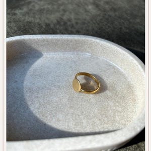 Op de afbeelding: Een goudkleurige ring met een cirkelvormige face. De ring ligt op een lichtgrijs, ovaalvormig dienblad. De ring heeft een dunne, gladde band en de face is een platte, effen cirkel.