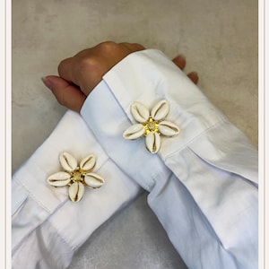 May include: A pair of flower-shaped cufflinks. Each cufflink features a cluster of white seashells arranged around a gold-colored center. The cufflinks are set against the backdrop of a white shirt sleeve.
