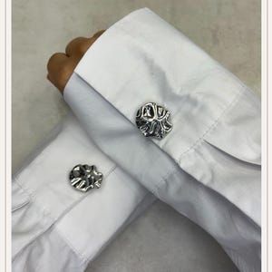 Puede incluir: Primer plano de unos gemelos plateados con un diseño abstracto y texturizado. Los gemelos están abrochados a los puños de una camisa blanca, mostrando los intrincados detalles de la joyería.