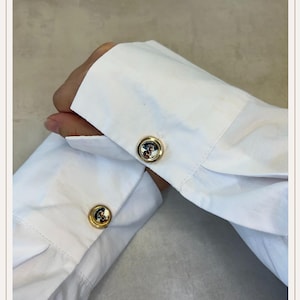 May include: Close-up of a white dress shirt with gold cufflinks. The cufflinks have a round design with a colorful image of a dog. The shirt sleeves are neatly cuffed, showcasing the cufflinks.