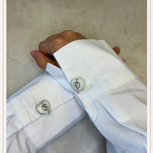 Op de afbeelding: Manchetknopen in hartvorm met een zilverkleurige zetting en een helder, iriserend midden. De manchetknopen zijn bevestigd aan de witte manchet van een shirt met lange mouwen. Close-up.
