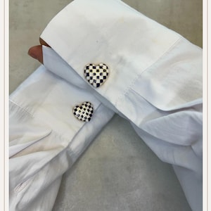 Peut inclure: Chemise blanche avec boutons de manchette en forme de cœur. Les boutons de manchette présentent un motif à carreaux noir et blanc avec une bordure bleu marine. La chemise est en tissu blanc.