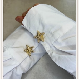 Op de afbeelding: Gouden zeester manchetknopen op een wit overhemd. De manchetknopen hebben een gestructureerd oppervlak en het overhemd heeft een klassieke button-down stijl. De manchetknopen voegen een nautisch thema toe aan de formele kleding.