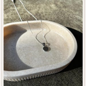 Op de afbeelding: Een zilveren ketting met een kleine, ronde hanger ligt in een lichtgekleurde, ovale schaal. De hanger is een eenvoudige, gepolijste schijf. De schaal heeft een textuur en staat tegen een neutrale achtergrond.