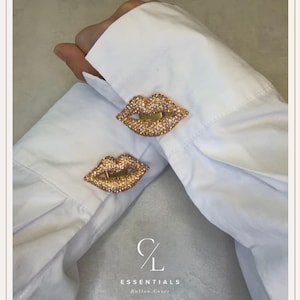 Può includere: Due copribottoni a forma di labbra decorati con piccoli cristalli scintillanti color oro rosa, fissati ai polsini di una camicia bianca a maniche lunghe. Il logo CL ESSENTIALS Button Cover è stampato in bianco nella parte inferiore.