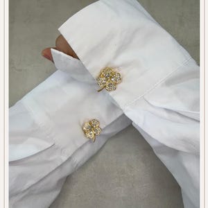 May include: Gold-coloured clover-shaped cufflinks with clear crystal detailing are fastened to the cuffs of a white dress shirt. The cufflinks have a detailed, three-dimensional design, adding elegance to the classic shirt.