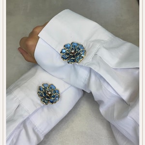Peut inclure: Boutons de manchette bleus en forme de fleur, avec une bordure dorée et un cristal central. Ils sont présentés sur les poignets d'une chemise blanche. Le design floral ajoute une touche d'élégance à une tenue formelle.