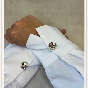Puede incluir: Gemelos plateados con un diseño de yin-yang colorido. Los gemelos redondos tienen un borde plateado y presentan un símbolo de yin-yang rosa, verde y negro. Se abrochan en una camisa blanca.