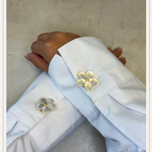 Può includere: Primo piano di polsini di camicia bianchi adornati da gemelli a forma di fiore. I gemelli presentano un design traslucido a forma di petalo con un centro dorato. La camicia è in tessuto bianco.