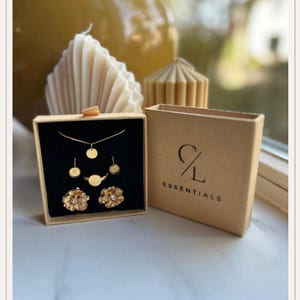 Op de afbeelding: Een goudkleurige sieradenset in een zwart gevoerde doos, inclusief een ketting, oorbellen, ring en bloemvormige oorstekers. Een bijpassende doos met de tekst "CL Essentials" staat naast de sieradendoos. Twee decoratieve kaarsen staan op de achtergrond.