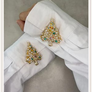 Pode incluir: Dois broches dourados em forma de árvore de Natal presos aos punhos de uma camisa branca de manga comprida. Cada alfinete em forma de árvore é decorado com pequenas pérolas brancas e strass multicoloridos em tons de vermelho, verde, azul e amarelo.