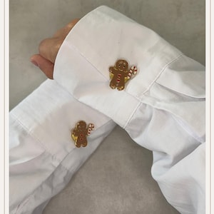 Op de afbeelding: Een paar manchetknopen van goudkleurig metaal in de vorm van peperkoekmannetjes, elk met een klein rood hartje op de borst en een rood-wit gestreepte zuurstok in de hand, bevestigd aan de manchetten van een wit overhemd met lange mouwen.