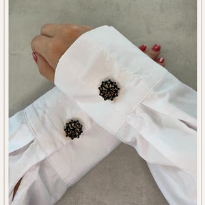 Pode incluir: Close-up de uma camisa branca com botões de punho pretos e dourados. Os botões de punho são redondos com um design floral e contrastam com o tecido branco das mangas.