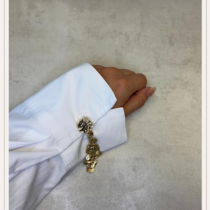 Può includere: Gemelli dorati con un design strutturato e organico. I gemelli sono attaccati a una camicia bianca a maniche lunghe. Il design dei gemelli presenta una parte superiore rotonda con una forma irregolare pendente.