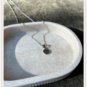 Op de afbeelding: Een zilveren ketting met een kleine, ronde hanger. De hanger is een effen, reflecterende cirkel. De fijne ketting ligt op een lichtgekleurd, getextureerd oppervlak. Het ontwerp is minimalistisch en elegant.