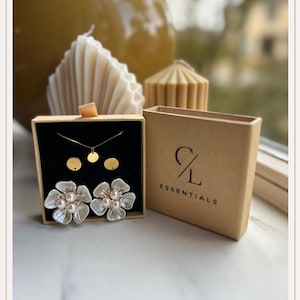 Op de afbeelding: Een sieradenset in een zwarte doos, met een gouden ketting met een kleine hanger, twee gouden oorstekers en een paar grote, witte bloemvormige oorbellen met parelcentra. Een bruine doos met de tekst "CL Essentials" is ook zichtbaar.