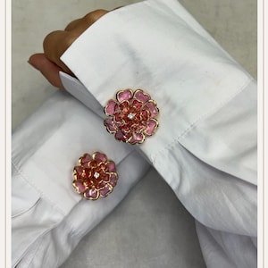 Peut inclure: Boutons de manchette en forme de fleur, avec une bordure dorée et un motif floral rose et rouge, chacun avec un petit bijou clair. Les boutons de manchette sont attachés à une chemise blanche.