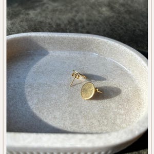 Op de afbeelding: Een paar goudkleurige oorstekers. Eén oorbel is een kleine, platte cirkel, de andere heeft een decoratief element. De oorbellen worden gepresenteerd op een lichtgrijze, ovaalvormige schaal.
