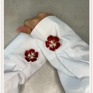 Può includere: Un paio di gemelli a forma di fiore rosso fissati ai polsini di una camicia bianca a maniche lunghe. Ogni gemello presenta cinque petali rossi arrotondati che circondano un gruppo centrale di tre piccole perle bianche su una base dorata.