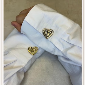 Peut inclure: Deux paires de boutons de manchette dorés en forme de cœur. Les boutons de manchette sont sur les poignets d'une chemise blanche boutonnée. Le design du cœur a un aspect texturé et organique.