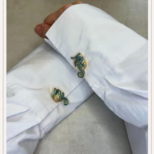 Pode incluir: Abotoaduras douradas com desenhos de cavalos-marinhos incrustados com pedras preciosas de cor turquesa. Os cavalos-marinhos têm uma cauda curva e uma cabeça pequena. As abotoaduras são colocadas numa camisa branca.