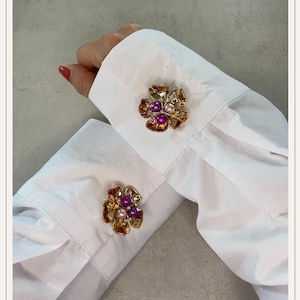 Peut inclure: Boutons de manchette dorés en forme de fleur avec des accents ambrés, violets et nacrés. Les boutons de manchette sont fixés aux poignets d'une chemise blanche. Le motif floral présente des pétales détaillés et un groupe central de petits ornements ronds.