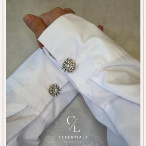 Pode incluir: Dois punhos de camisa brancos com capas de botão circulares em tom prateado, adornadas com cristais claros e brilhantes dispostos num padrão de raios de sol. O logótipo CL ESSENTIALS Button Cover está impresso a branco na base da imagem.