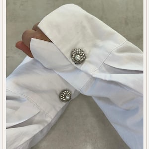 Può includere: Primo piano di una camicia bianca con gemelli decorativi. I gemelli sono color argento, con una perla centrale circondata da piccoli cristalli scintillanti. Il tessuto della camicia è fresco e pulito.