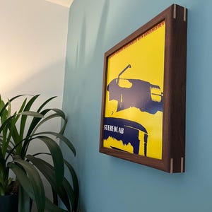 May include: A framed record album with a yellow background and a blue illustration of a record player. The album title is "The  Sound of  Vinyl  Noise Bursts with Announcement" and the artist is "Stereolab".