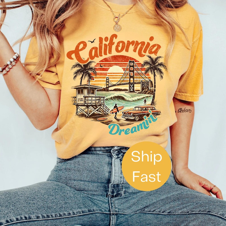 May include: Mustard yellow t-shirt with a graphic featuring the word "California" in a retro font, a sunset, palm trees, the Golden Gate Bridge, and the word "Dreamin".