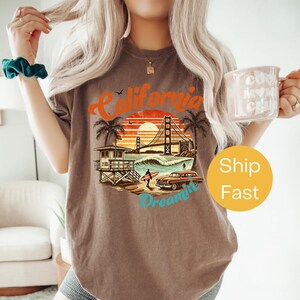 May include: Brown t-shirt with a California graphic featuring the Golden Gate Bridge, a surfer, and a vintage car. The design includes the words "California Dreamin'" in a retro style. The shirt is a relaxed fit.